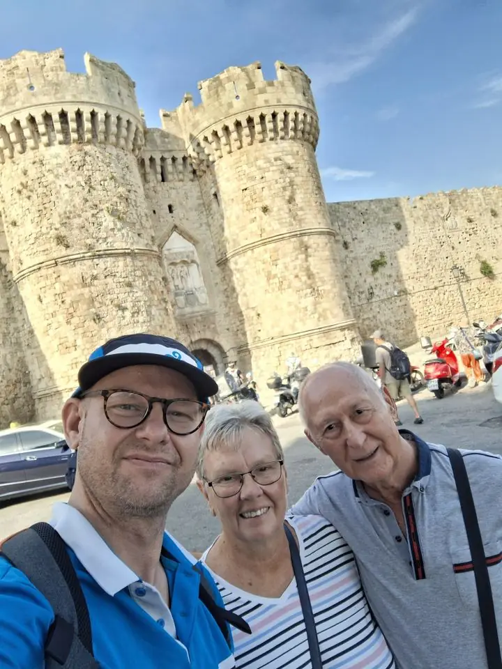 A man in a cap and glasses, a smiling woman and an older man taking a selfie in front of the massive stone towers of a medieval fortress gate in Rhodes