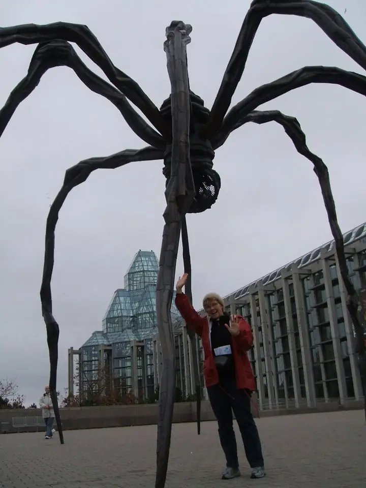 A smiling person in a red jacket waving beneath a giant black spider sculpture outside a glass-towered gallery in Ottawa
