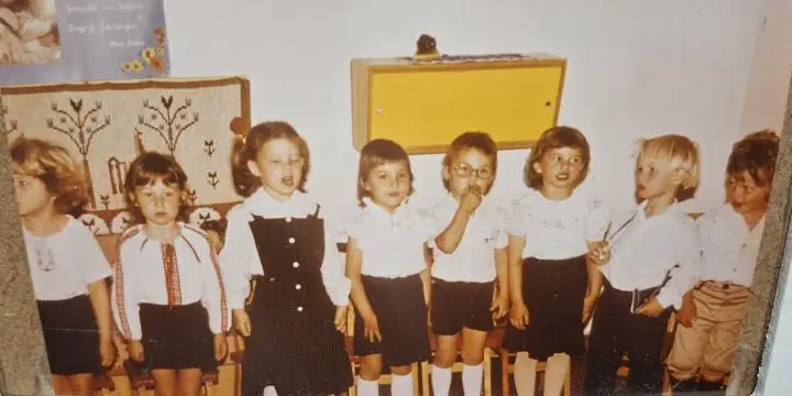 A row of small kindergarten children in white shirts and dark shorts and skirts standing in front of a wall hanging