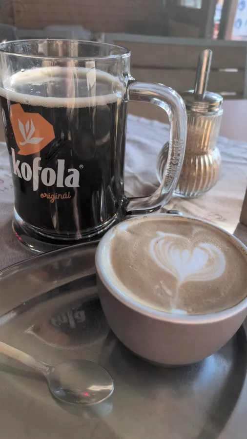 A cappuccino with a latte-art heart next to a mug of Kofola, at Komp Café in Štúrovo