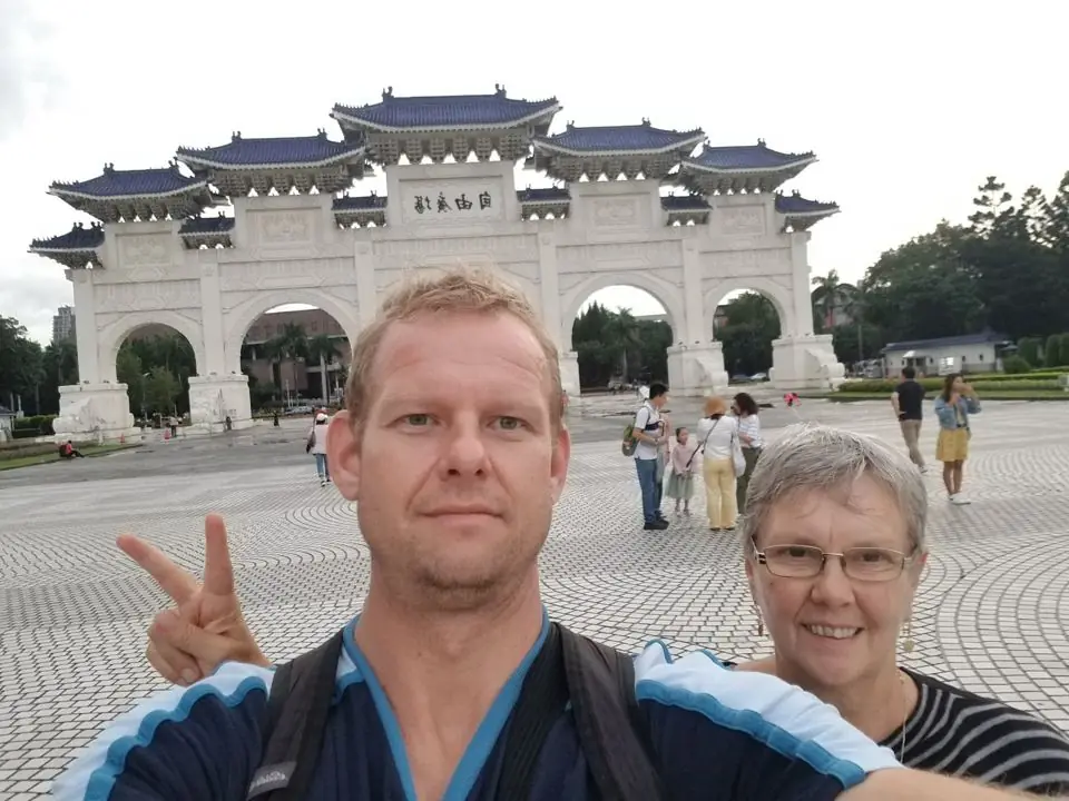 A man making a peace sign and his mother taking a selfie in front of a large white gate with blue tiled roofs on a wide plaza in Taipei