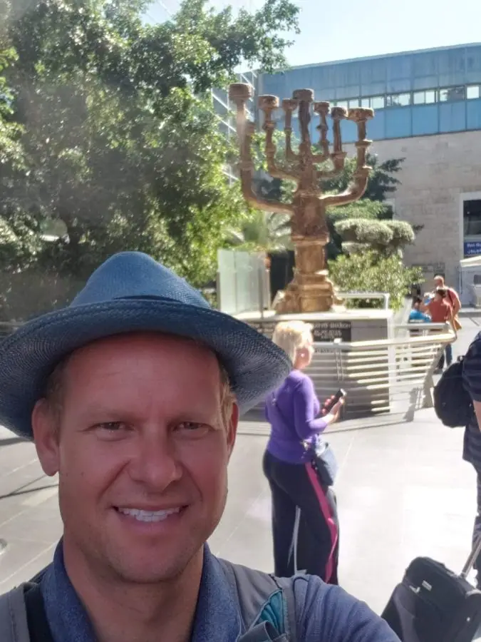 A smiling man in a blue hat taking a selfie in front of a large bronze menorah sculpture in a plaza